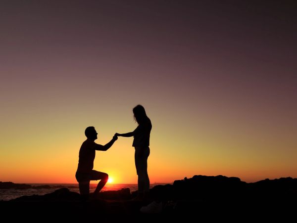 Silhouette of a man proposing to a woman at sunset by the water.