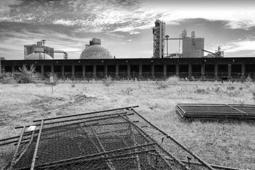 Black and white photo of industrial site and buildings with fence in foreground.