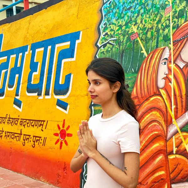 A woman in white performs a prayer gesture near colorful wall art.