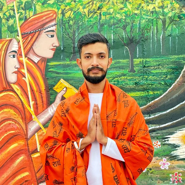 Man in orange robe with hands folded in prayer in front of a mural.