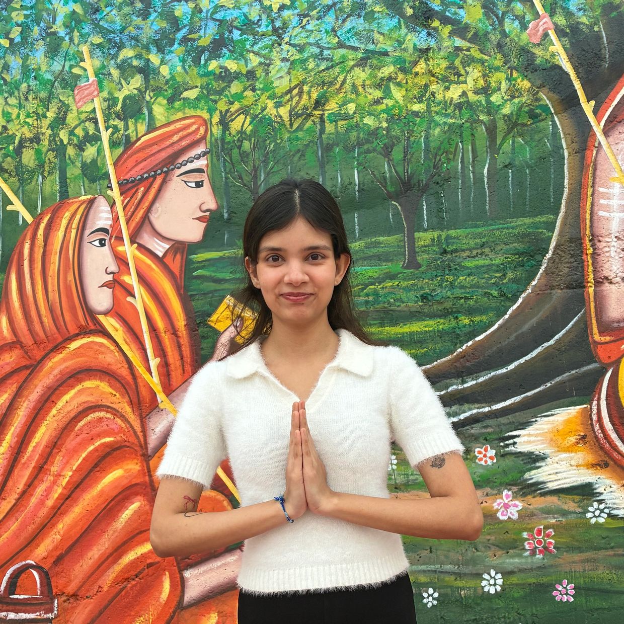 Young woman posing with namaste gesture in front of colorful mural.