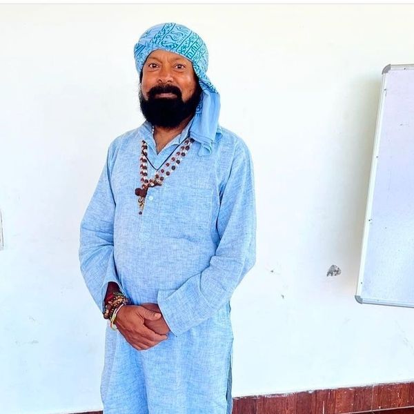 Man wearing a blue traditional outfit and headscarf, standing indoors.