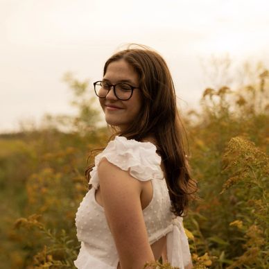 Young woman in glasses smiles softly in a field of tall plants during golden hour.