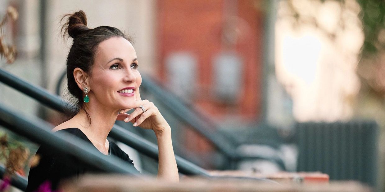A woman with a thoughtful smile sitting outdoors, resting her chin on her hand.