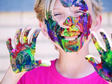 Child with colorful paint on face and hands smiling joyfully.