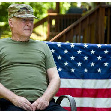 An elderly man in military attire sitting with an American flag behind him.