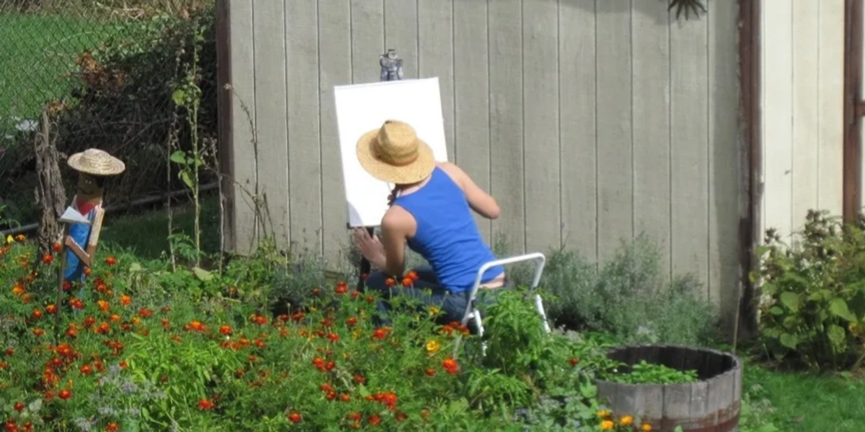 Artist sitting in a garden in front of an easel. Artist is wearing a wide brim hat. 