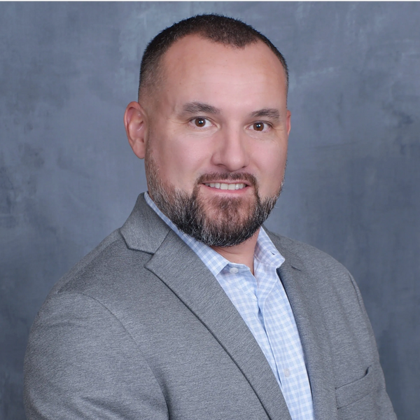 Professional headshot of a smiling man in a gray blazer and checkered shirt.