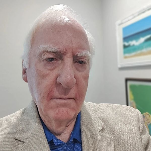 Elderly man in beige blazer and blue shirt with serious expression indoors.