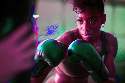 A young black woman in green boxing gloves gets ready to punch a heavy bag.