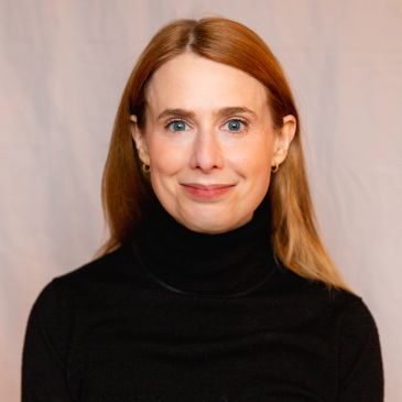 Portrait of a woman with red hair wearing a black turtleneck, smiling gently.