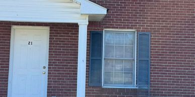 Brick house exterior with white door numbered 21 and window with blue shutters.