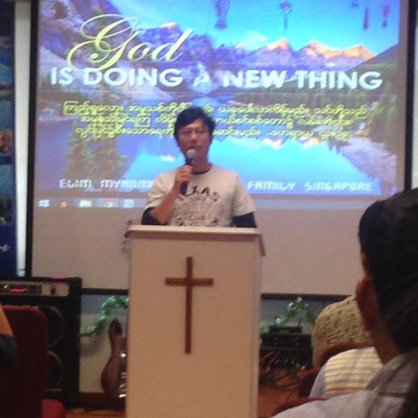 Man speaking at a church podium with a cross and a religious message on screen.