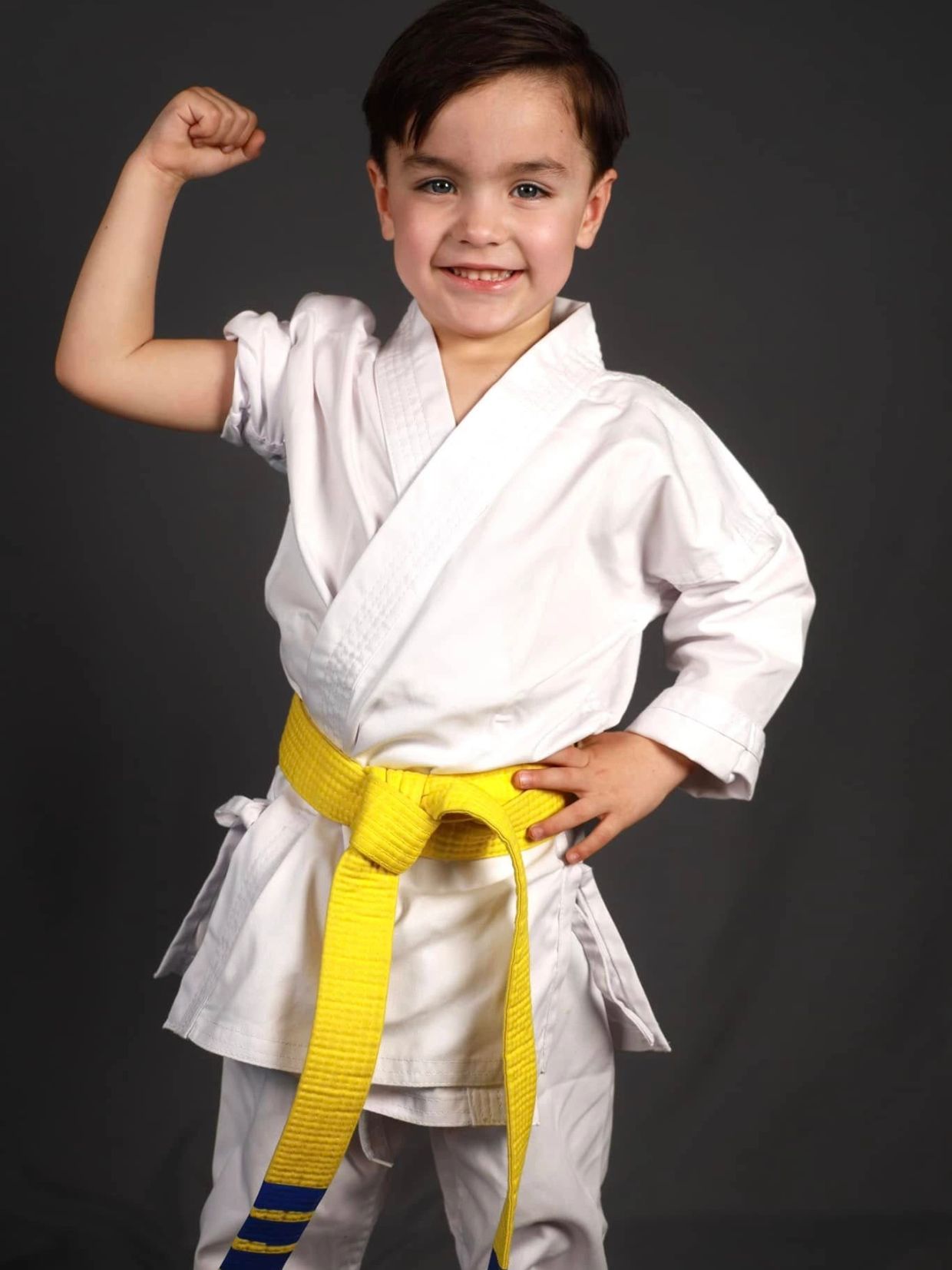Young karate student wearing a white uniform and yellow belt, smiling and flexing with confidence at West Coast Karate.