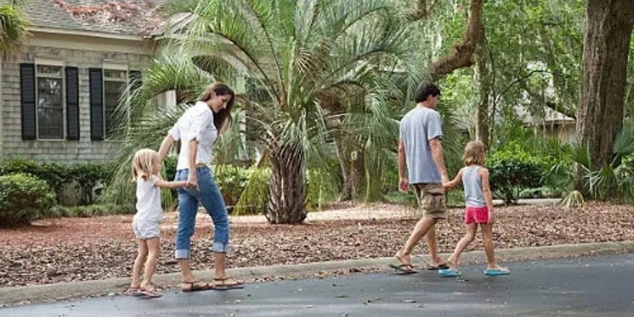 A family of four walking hand in hand outdoors near a house with lush greenery.