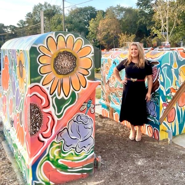 Woman in black dress standing beside colorful flower-painted utility boxes outdoors.