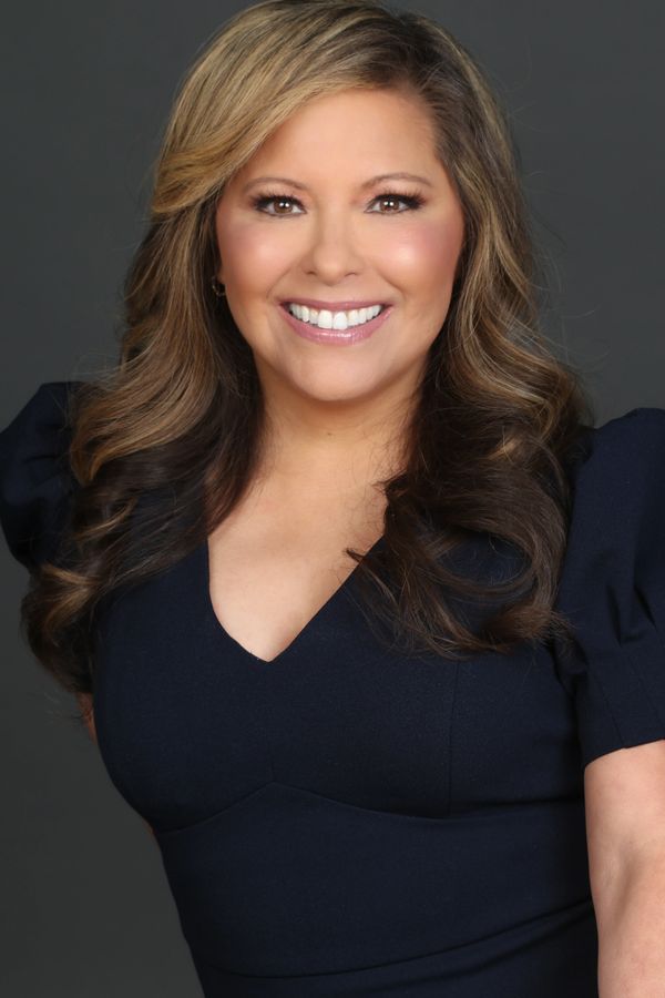 Smiling woman with wavy highlighted hair and a navy blue dress against a dark background.