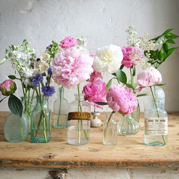 Vintage glass bottles hold delicate pink and white peonies and wildflowers on a rustic wooden table.
