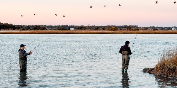 Two fishermen standing in water fishing during sunset with birds flying overhead.