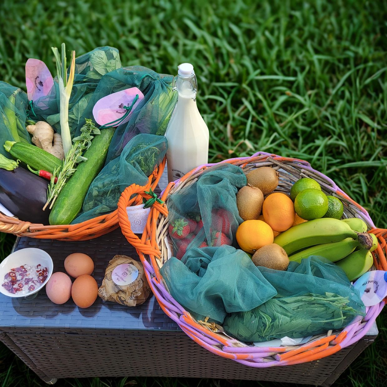 Two baskets of fresh fruits, vegetables, eggs, and a milk bottle on a table outdoors.