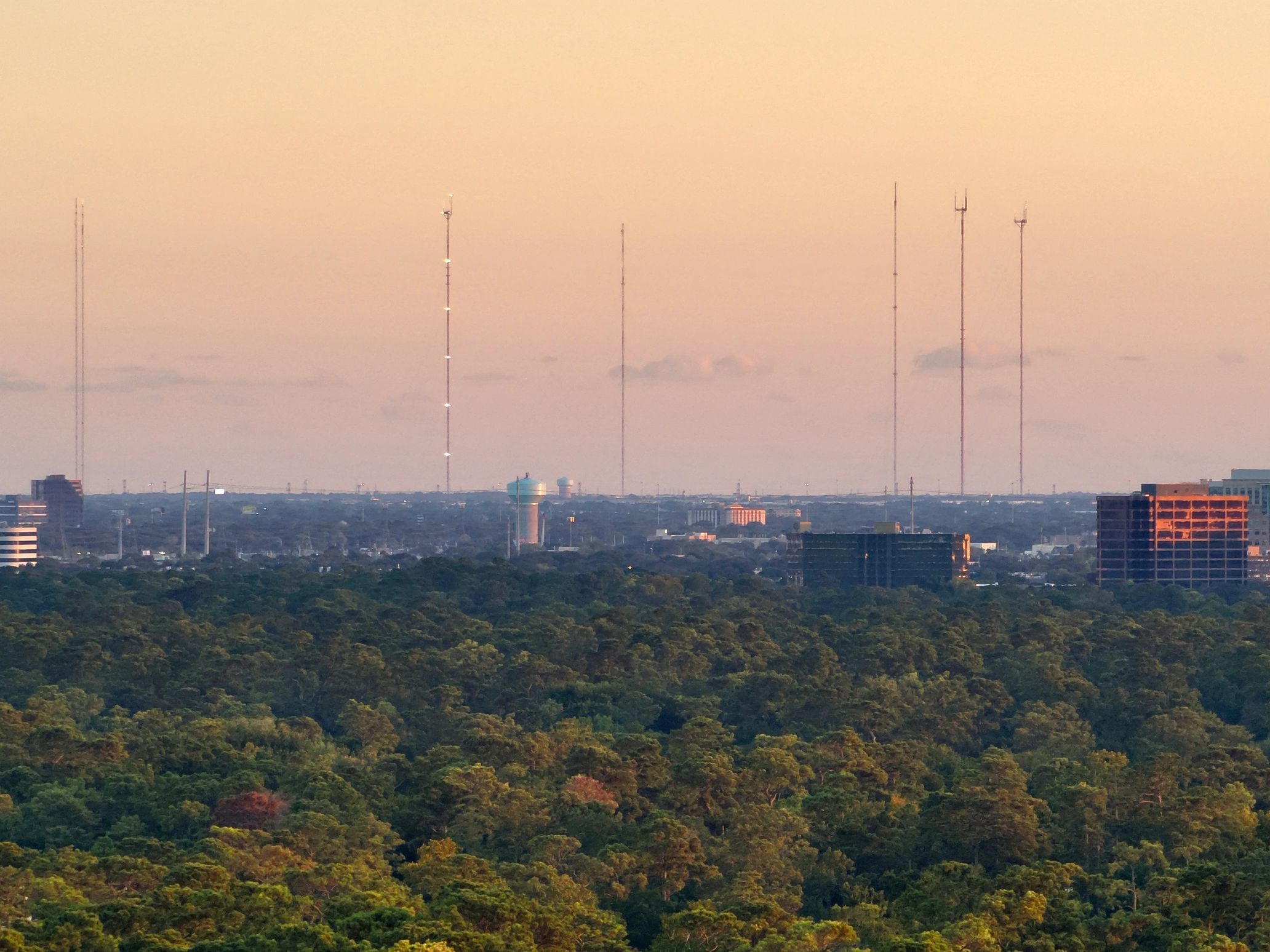 City skyline with tall antennas and lush green forest at sunset.
