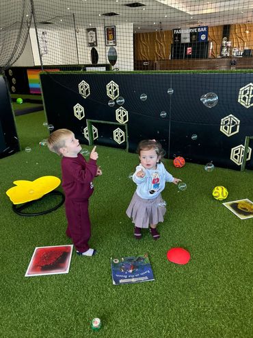 Two toddlers play with bubbles on artificial grass indoors.