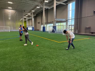 Three people stretching on an indoor soccer field with goals and cones.