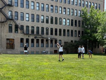 People playing catch on a grassy field in front of a large brick building.