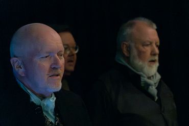 Three older men in dark clothing and scarves singing or speaking in a dimly lit setting.