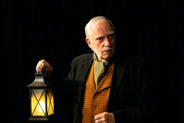 Elderly man holding a glowing lantern against a dark background.