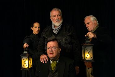 Four people in dark clothing hold lanterns in a dramatic, dimly lit setting.