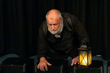A man with a beard leans forward, hands on black boxes, beside a glowing lantern.