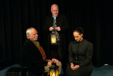 Three actors in Victorian attire perform a dramatic scene with lanterns.