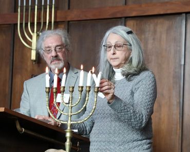 Two elderly people lighting candles on a menorah inside a synagogue.