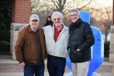 Three older men smiling outdoors with a blue inflatable dreidel in the background.