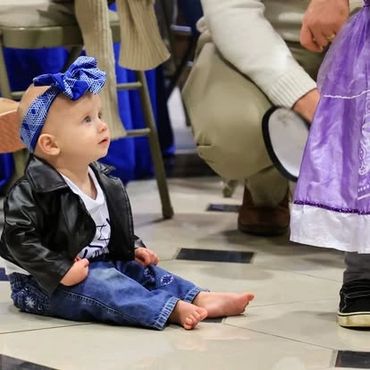 Baby girl in a leather jacket and headband sitting barefoot on the floor.