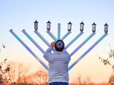 Person lighting a large menorah outdoors at sunset.