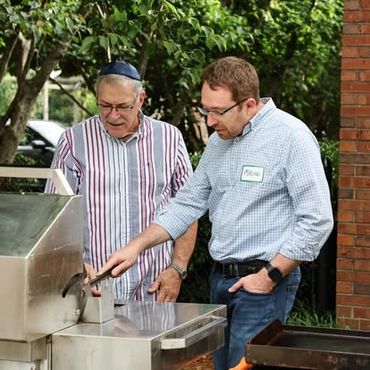 Two men grilling outdoors, one pointing at the grill.