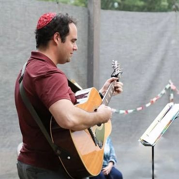 Man playing acoustic guitar outdoors, wearing a red kippah.