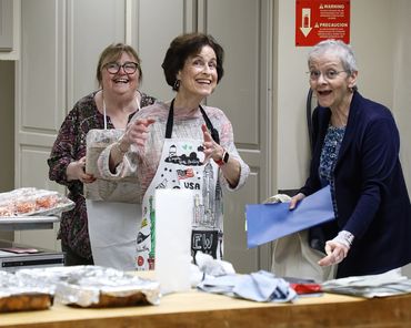 Three joyful women in aprons preparing food in a kitchen.