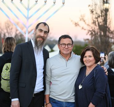 Three people posing together outdoors with a large menorah in the background.