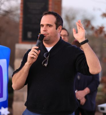 Man speaking into a microphone outdoors, gesturing with one hand raised.