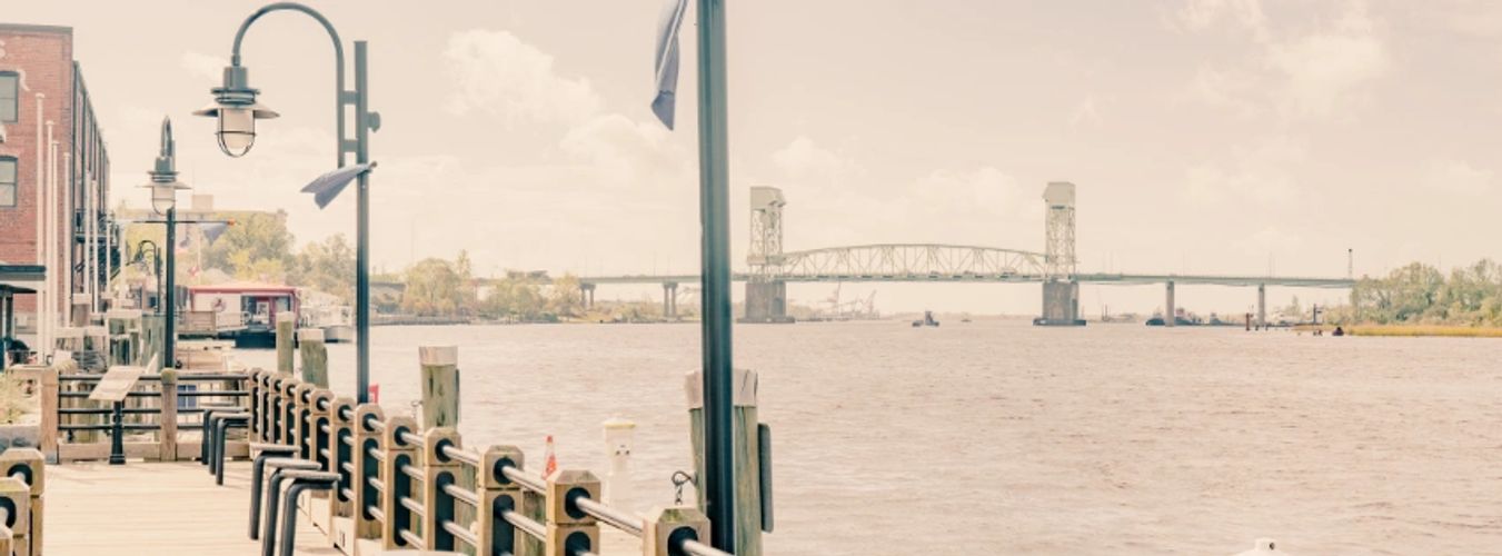 Calm riverside boardwalk with benches and a distant bridge under a soft sky.