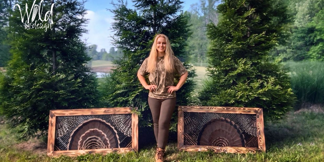 Young woman standing between two wooden turkey feather frames in a forest clearing.