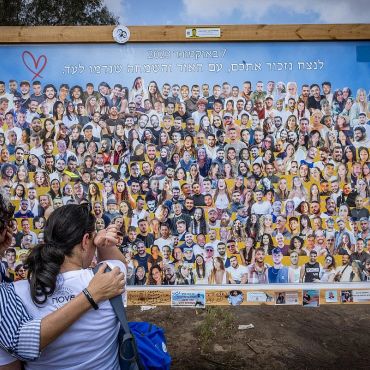 Two people mourning in front of a memorial collage of many faces.
