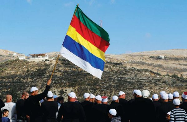 A group of people, many wearing white caps, with one person holding a colorful striped flag outdoors.