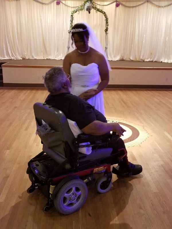 Bride in white gown lovingly holding hands with an elderly man in a wheelchair.