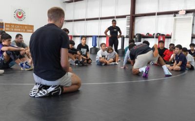 A group of athletes sits on the edge of a wrestling mat inside a gym, watching a coach demonstrate a