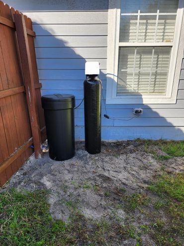 Black water softener system installed beside a window and wooden fence.