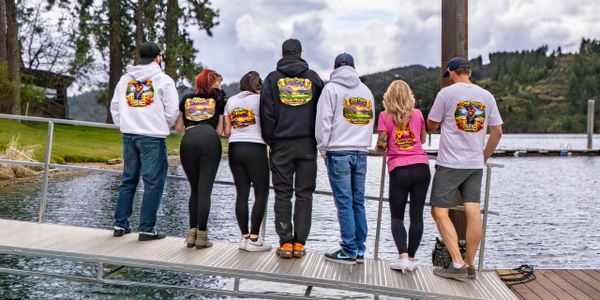 Seven people stand on a dock, showing colorful designs on their hoodies and t-shirts.